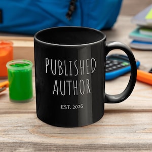 May include: A black coffee mug with the words "PUBLISHED AUTHOR" and "EST. 2026" in white. The mug is on a wooden surface with art supplies and a blue backpack in the background. The mug is a perfect gift for writers.