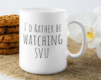 May include: White ceramic mug with the text "I'd rather be watching SVU" in black lettering. A stack of chocolate chip cookies and a white flower are next to the mug. The mug has a curved handle.