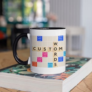 May include: White ceramic mug with a black handle. The mug features a Scrabble board design with the word "CUSTOM WORDS" spelled out in colourful tiles.
