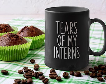 May include: A black coffee mug with the text "TEARS OF MY INTERNS" in white. The mug is next to three chocolate muffins in green paper cups and scattered coffee beans on a green and white checkered cloth.