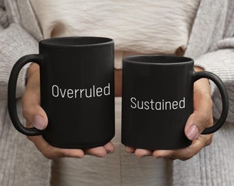 May include: Two black ceramic mugs with white text. One mug reads "Overruled" and the other reads "Sustained". The mugs have a curved handle and are held up in front of a person.