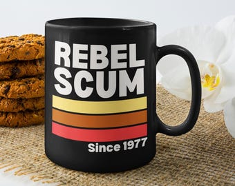 May include: Black ceramic mug with the text "REBEL SCUM" in white, above horizontal stripes of yellow, brown, and red. The text "Since 1977" is below the stripes. A stack of chocolate chip cookies is to the left.