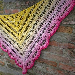 Puede incluir: Un chal tejido a mano, con un degradado de colores. El chal pasa del amarillo al rosa, con un borde festoneado en un tono rosa vibrante. El diseño calado añade un toque delicado a este accesorio.