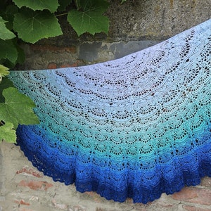 Könnte beinhalten: Ein gehäkeltes Tuch in Blautönen, Grün und Weiß mit einem gewellten Rand. Das Tuch ist über eine Backsteinmauer gelegt.