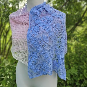 May include: Two crocheted shawls, one in light pink and the other in light blue, displayed on a mannequin. The shawls have a delicate, lacy pattern. The background is a blurred view of green foliage.