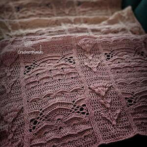 Puede incluir: Primer plano de una manta de ganchillo hecha a mano en tonos malva y crema. La manta presenta intrincados calados y patrones texturizados, incluyendo diseños en forma de abanico y motivos florales. El texto "crochethands" es visible.