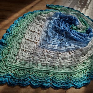 May include: A handmade, crocheted shawl in shades of blue, green, and white. The shawl features a spiral design with a scalloped edge. The colors transition from a deep blue center to lighter shades of green and white at the edges. The shawl is made of soft yarn.