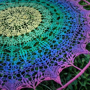 May include: A handmade, circular dream catcher with a vibrant, rainbow-colored crochet design. The crochet pattern transitions from yellow to green, blue, and purple, with a pink outer ring. The dream catcher is set against a backdrop of green grass.
