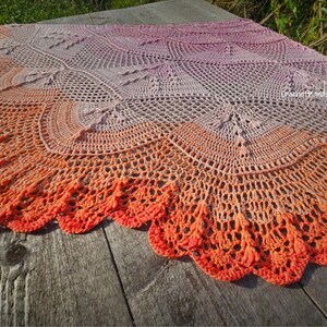 May include: A handmade crochet shawl featuring an intricate geometric pattern with a gradient color transition from light lavender at the center to a vibrant orange at the scalloped edge. The textile includes the text CrochetHands in white.