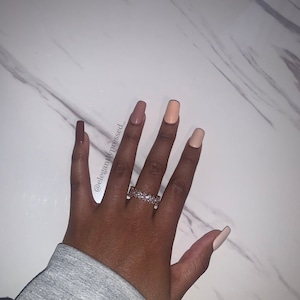 May include: A hand with nude, light brown, and brown nail polish. The hand is wearing a ring with small clear stones.