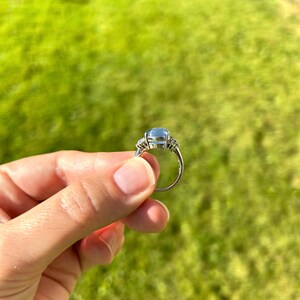 May include: A silver ring with a large, pale blue gemstone and smaller, clear stones. The ring is held up against a green grassy background, highlighting its elegant design and the sparkle of the gems.