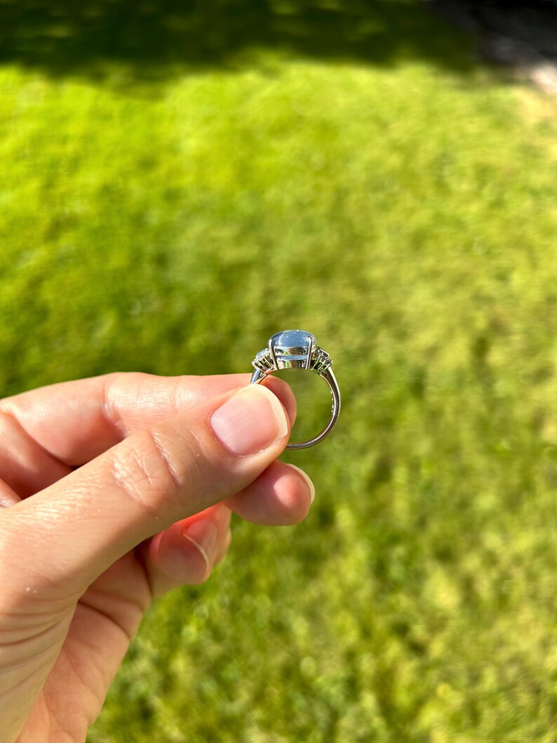 May include: A silver ring with a large, pale blue gemstone and smaller, clear stones. The ring is held up against a green grassy background, highlighting its elegant design and the sparkle of the gems.
