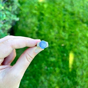 May include: A silver ring featuring a large, oval, pale blue gemstone. The ring is flanked by smaller, clear stones. The ring is held up against a backdrop of green grass.