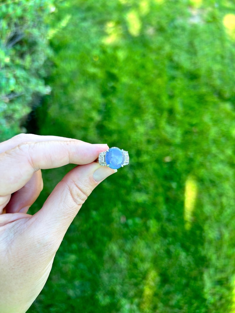 May include: A silver ring featuring a large, oval, pale blue gemstone. The ring is flanked by smaller, clear stones. The ring is held up against a backdrop of green grass.