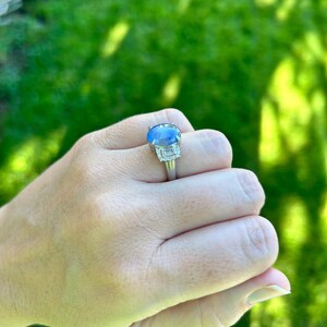 May include: A silver ring with a large, oval blue gemstone at its centre. Two smaller, rectangular clear gemstones are set on either side. The ring is displayed against a green background.