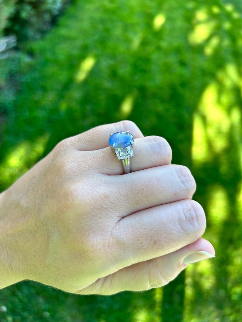 May include: A silver ring with a large, oval blue gemstone at its centre. Two smaller, rectangular clear gemstones are set on either side. The ring is displayed against a green background.