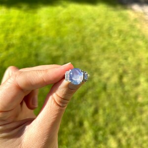 May include: A silver ring with a large, oval, pale blue gemstone. The ring has smaller, rectangular stones flanking the main stone. The ring is held up against a backdrop of green grass.