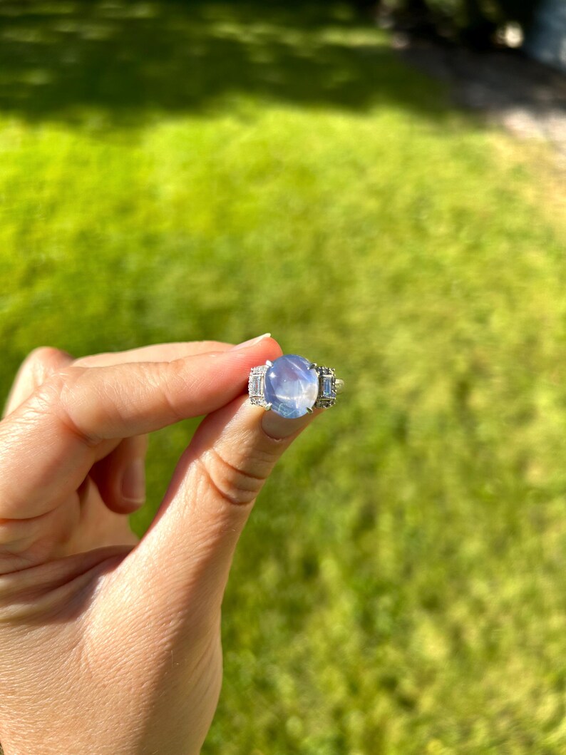 May include: A silver ring with a large, oval, pale blue gemstone. The ring has smaller, rectangular stones flanking the main stone. The ring is held up against a backdrop of green grass.