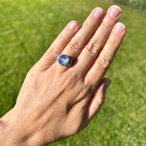 May include: A silver ring featuring a large, round, pale blue gemstone. The ring has a detailed setting with smaller, clear stones. The ring is shown on a hand against a backdrop of green grass.