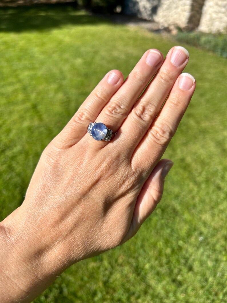 May include: A silver ring featuring a large, round, pale blue gemstone. The ring has a detailed setting with smaller, clear stones. The ring is shown on a hand against a backdrop of green grass.
