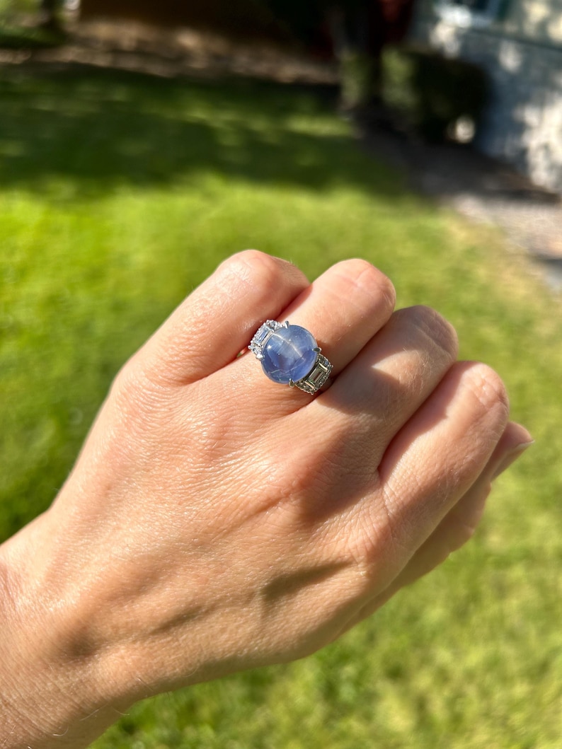May include: A silver ring with a large, round, pale blue gemstone as the focal point. The ring has smaller, rectangular, clear stones flanking the main stone. The ring is on a hand, with a green background.
