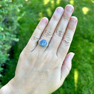 May include: A silver ring with a large, oval, light blue gemstone is worn on a hand. The ring features smaller, clear stones flanking the main stone. The background is a blurred green lawn.