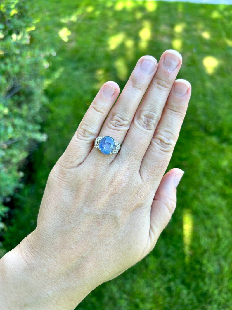 May include: A silver ring with a large, oval, light blue gemstone is worn on a hand. The ring features smaller, clear stones flanking the main stone. The background is a blurred green lawn.