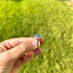 May include: A silver ring featuring a large, oval-cut, pale blue gemstone. The gemstone is encircled by a halo of smaller, clear stones. The ring is held against a backdrop of green grass.