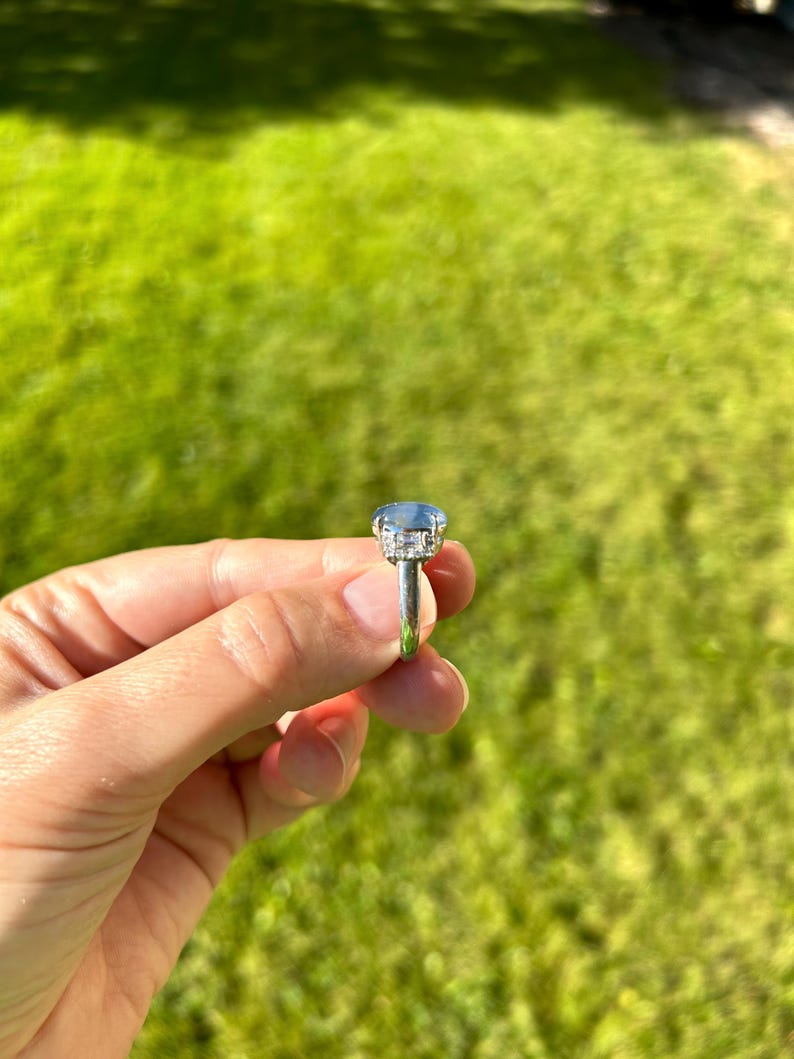 May include: A silver ring featuring a large, oval-cut, pale blue gemstone. The gemstone is encircled by a halo of smaller, clear stones. The ring is held against a backdrop of green grass.