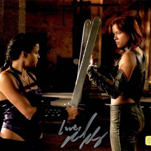 May include: Two women in a dimly lit room, one holding two silver swords. They are dressed in dark, form-fitting outfits. The woman on the left is wearing a purple bodysuit, while the other is in black leather pants and a top. A signature is visible.