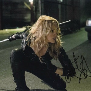 May include: A woman in a black leather jacket and pants holds a silver staff in a fighting stance. She has long blonde hair and is looking to the right. The background is a dark alley.
