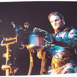 May include: A man in a futuristic, dark blue and gray suit holds a large, silver, futuristic weapon. He is looking directly at the camera. The weapon has a large, silver barrel and a black handle. The man is wearing a black belt with a silver buckle. The background is dark.