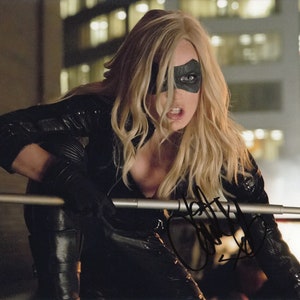 May include: A woman wearing a black leather jacket and pants with a black eye mask. She is holding a silver metal staff. The background is a blurry city scene.