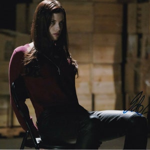 May include: A woman with long dark hair is sitting on a black folding chair. She is wearing a maroon long-sleeved shirt and black pants. The background is a wall of wooden crates.