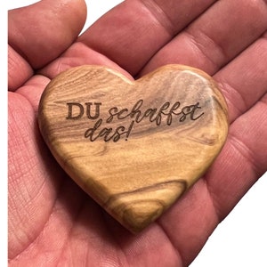 Könnte beinhalten: Ein herzförmiges Holz-Andenken mit dem eingravierten Text "DU schaffst das!". Das Holz hat eine hellbraune Farbe mit dunkleren Maserungsmustern. Das Herz wird in einer offenen Hand gehalten.