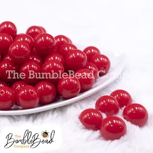 May include: A pile of bright red round beads in a white bowl. The beads are smooth and shiny. There are also a few red beads scattered on a white fuzzy surface.