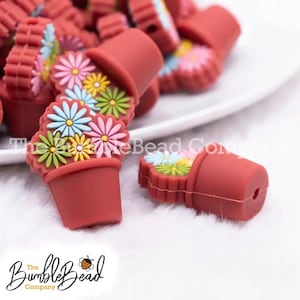 May include: A close-up of red flower pot shaped silicone beads with colorful flower designs. The beads are scattered on a white surface.