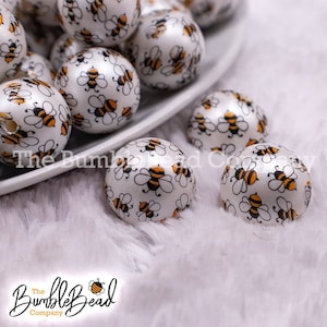 May include: White round beads with a repeating pattern of black and orange bees. The beads are on a white surface and some are in a white dish. The BumbleBead Company logo is in the bottom left corner.