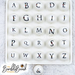 May include: White round beads with black letters A through Z and an ampersand. The beads are arranged in a grid on a white surface. The text "The BumbleBead Company" is printed below the grid.