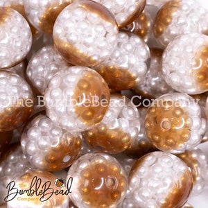 May include: A close-up of a group of round, clear, plastic beads with a white pearl-like finish and a brown ombre effect. The beads are scattered on a surface.