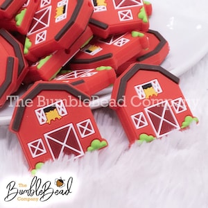 May include: A close-up of red silicone barn charm beads with brown roofs, white windows, and yellow hay sticking out of the windows. The barn charms have a red door with white X's and green grass at the bottom.