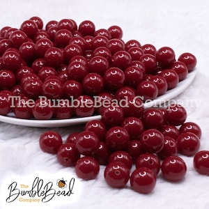 May include: A large pile of 20mm red round acrylic beads. The beads are glossy and have a smooth finish. The beads are on a white plate and a white fuzzy surface.