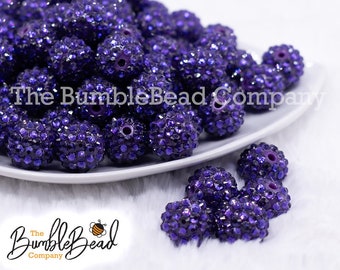 May include: A close-up of a white plate filled with purple rhinestone beads. The beads are round and have a sparkly finish. The plate is sitting on a white fluffy surface. The text "The Bumble Bead Company" is visible in the background.