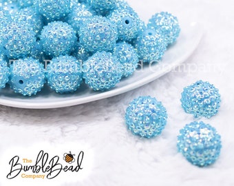 May include: A pile of light blue, sparkly, round beads on a white plate.  The beads have a textured surface and are scattered on a white fluffy surface.  The Bumble Bead Company logo is in the bottom left corner of the image.