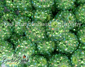 Pode incluir: Close-up de uma pilha de contas verdes cobertas de strass. As contas são redondas com um buraco no centro. O logotipo da The BumbleBead Company é visível.