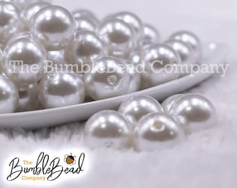 May include: A white bowl filled with white pearl beads. The beads are round and smooth, with a slight shine. The bowl is sitting on a white fluffy surface.