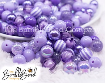 May include: A large assortment of purple and lavender colored beads. The beads are made of various materials, including plastic, glass, and acrylic. Some beads have a glitter finish, while others have a smooth, matte finish. The beads are arranged in a pile on a white surface.