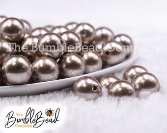 May include: A pile of large, round, pearl-like beads in a champagne color. The beads are arranged on a white plate and some are scattered on a white fluffy surface. The beads have a smooth, glossy finish.