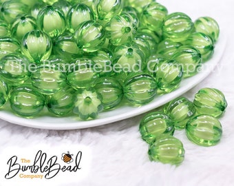 May include: A close-up of a bowl filled with green, translucent, faceted beads. The beads are oval-shaped and have a ribbed texture. The beads are arranged in a random pattern and are scattered on a white surface.