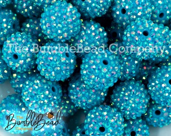 May include: A close-up of a pile of turquoise blue round beads covered in iridescent rhinestones. The beads have a hole in the center for stringing.